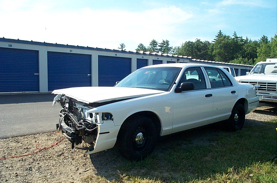 White sedan with front-end damage parked in front of storage units. Another white truck is parked to the right
