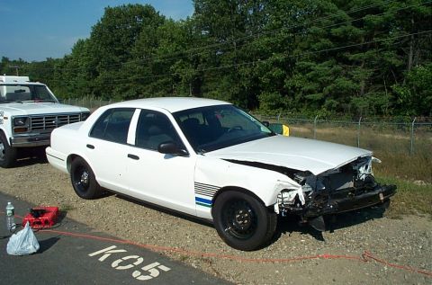 2001 Ford Crown Victoria - A damaged white 2001 Ford Crown Victoria is parked beside a tow truck on a gravel lot.