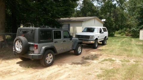 1996 Ford Bronco - Two SUVs, a 1996 Ford Bronco and a Jeep Wrangler, are parked outside a building.