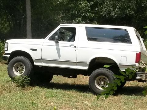 1996 Ford Bronco - A white 1996 Ford Bronco is shown from the side, parked in a grassy area with trees in the background.