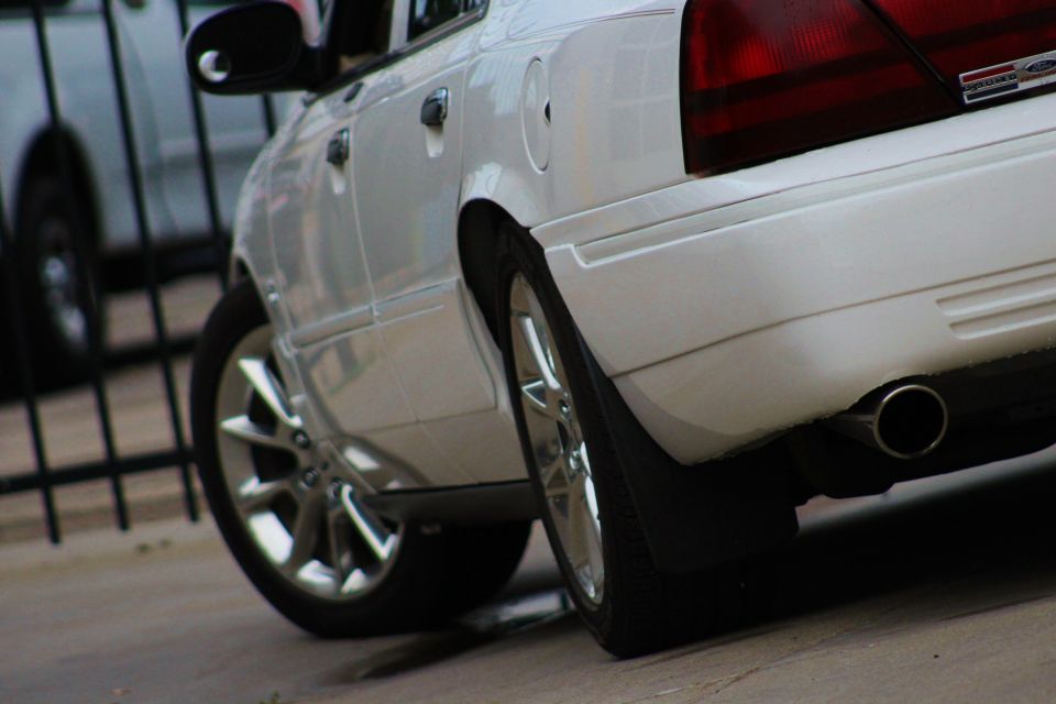 White sedan's rear quarter panel and wheel. Another car is visible behind a fence