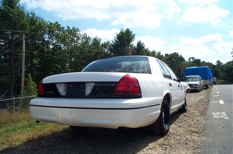 2001 Ford Crown Victoria - A white 2001 Ford Crown Victoria is seen from the rear, parked on the side of a road with other vehicles in the background.