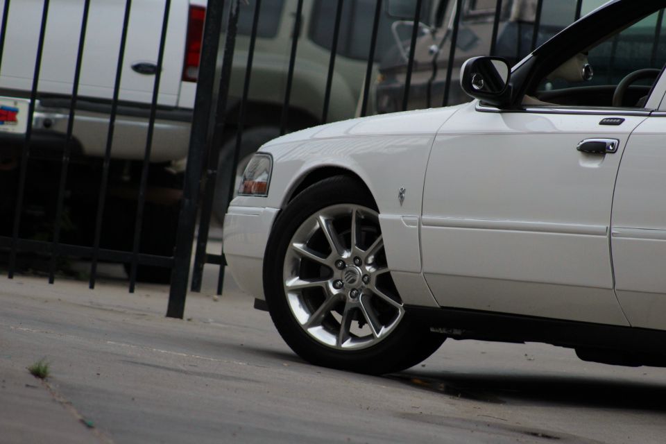 White sedan front quarter panel and wheel, parked behind a black fence. Another white vehicle is visible behind the fence
