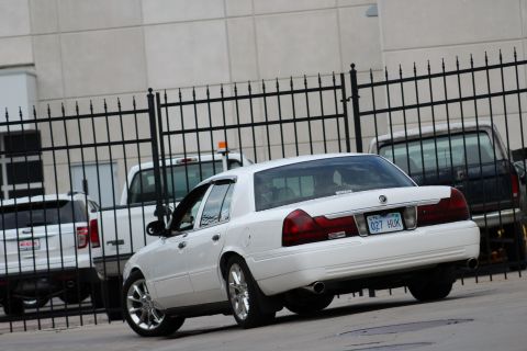 2000 Mercury Grand Marquis - A white 2000 Mercury Grand Marquis is parked behind a fence, showing its rear profile and custom wheels.