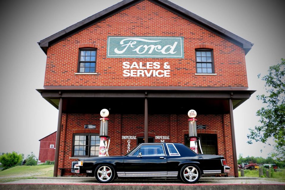 Black 1980s Ford sedan parked in front of a brick building with a Ford Sales & Service sign and vintage gas pumps