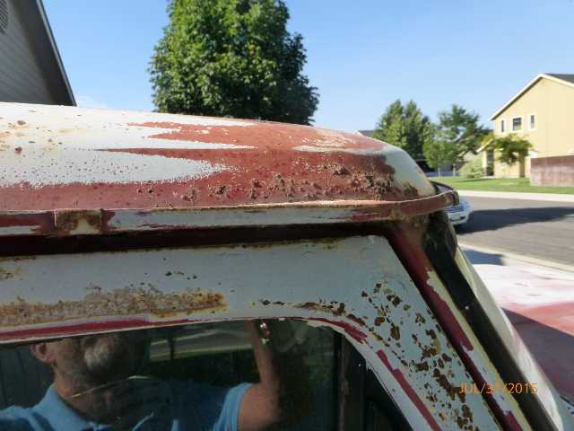 Close-up of a rusty white and red truck cab corner, showing peeling paint and rust damage. A man's reflection is visible in the window