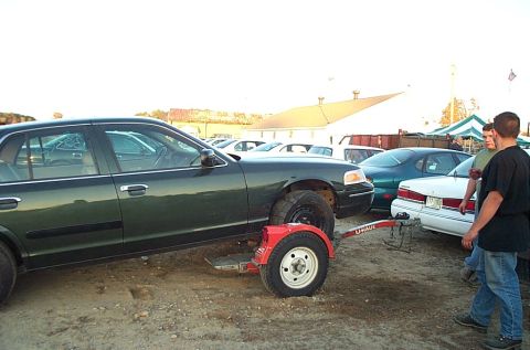 2001 Ford Crown Victoria - A dark green 2001 Ford Crown Victoria is loaded onto a U-Haul dolly, with a flat tire visible on the dolly's wheel.