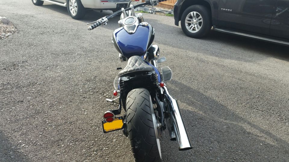 Rear view of dark blue cruiser motorcycle with wide rear tire and chrome exhaust on gravel driveway