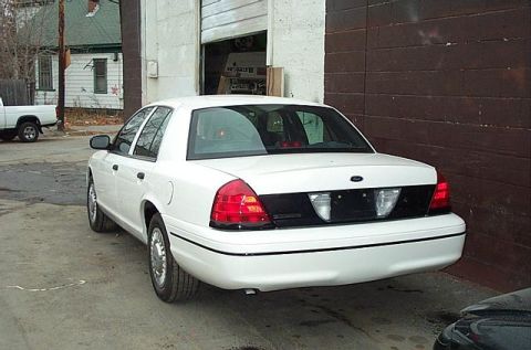 2001 Ford Crown Victoria - The rear of a white sedan is parked outdoors near a garage. A white pickup truck is visible in the background.