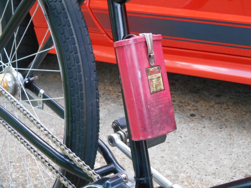 Close-up of bicycle rear wheel, chain, and red carbide lamp, with a red vehicle in the background.