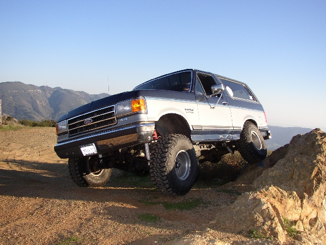 Lifted silver and blue Ford Bronco SUV climbing a rock on a dirt trail with mountains behind.