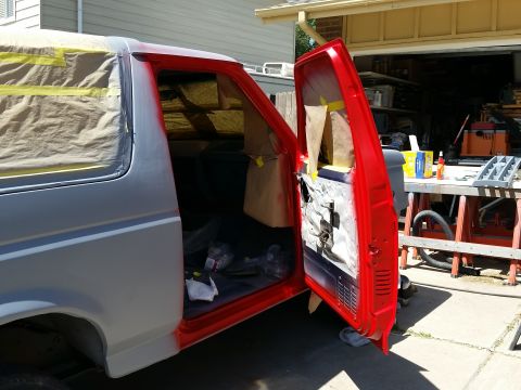 1988 Ford Bronco - A red-painted driver's side door of a Ford Bronco is open, revealing the interior and the vehicle's gray body. The vehicle is parked outdoo…