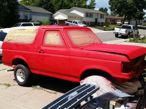 1988 Ford Bronco - A red Ford Bronco is being prepped for paint, with masking tape covering the windows and a textured red spray coating applied to the body.…