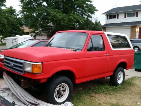 1988 Ford Bronco - A red 1988 Ford Bronco with a white top is parked outdoors next to a house and some trees. The vehicle appears to be undergoing some restor…