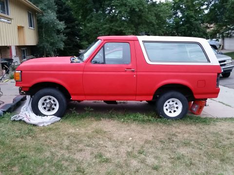 1988 Ford Bronco - The side profile of a red Ford Bronco is shown, with a white roof and white rims. The vehicle is parked on grass next to a driveway, with a…