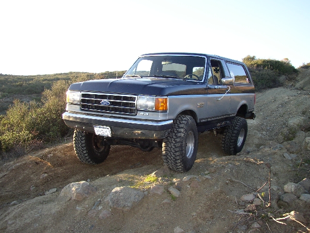 Blue and silver lifted Ford Bronco SUV on off-road dirt trail, front 3/4 view