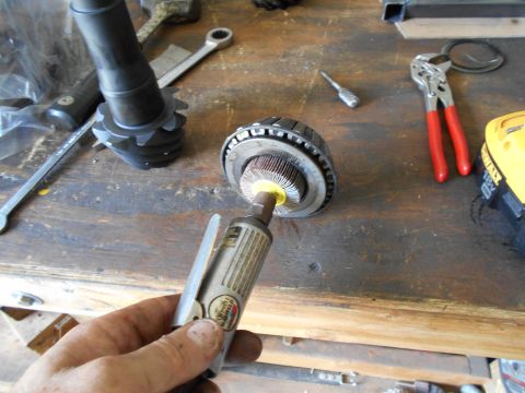 1992 Ford Bronco - A hand holds a pneumatic grinder with a Scotch-Brite wheel attached, positioned over a differential gear assembly on a wooden workbench. Va…
