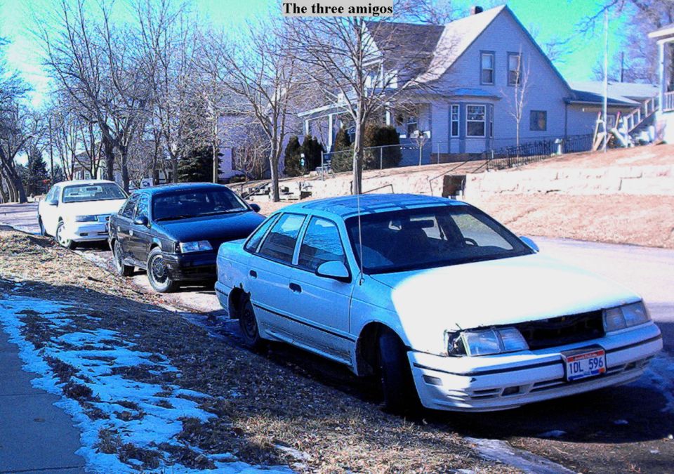 White sedan parked at curb with two more cars behind, near a blue house with snow patches.