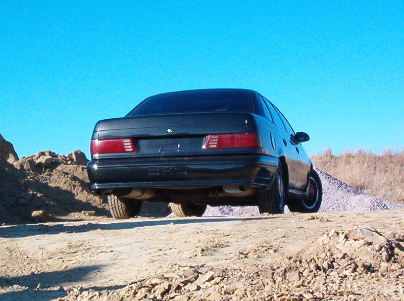 Rear view of a dark green Ford Taurus sedan on a dirt embankment under blue sky