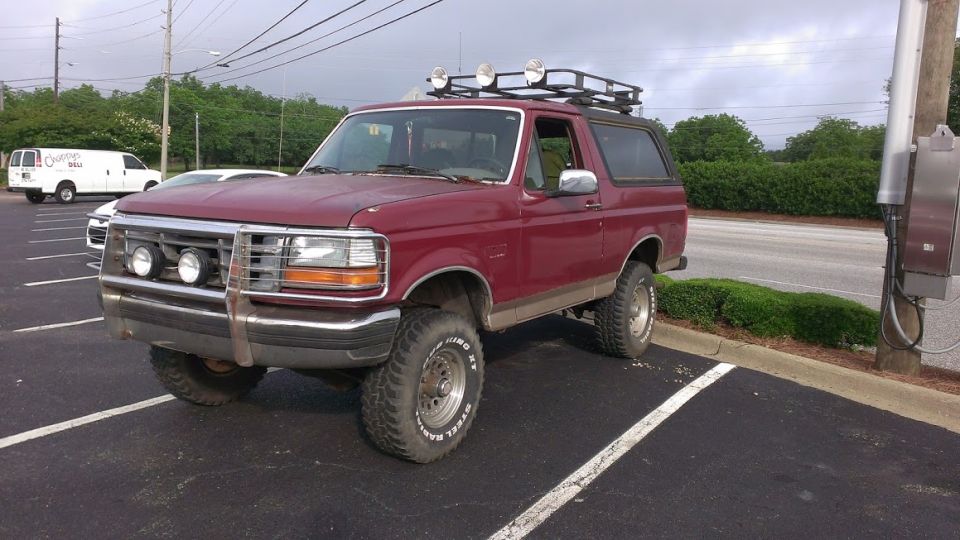 Maroon Ford Bronco with roof rack and brush guard parked in a lot.