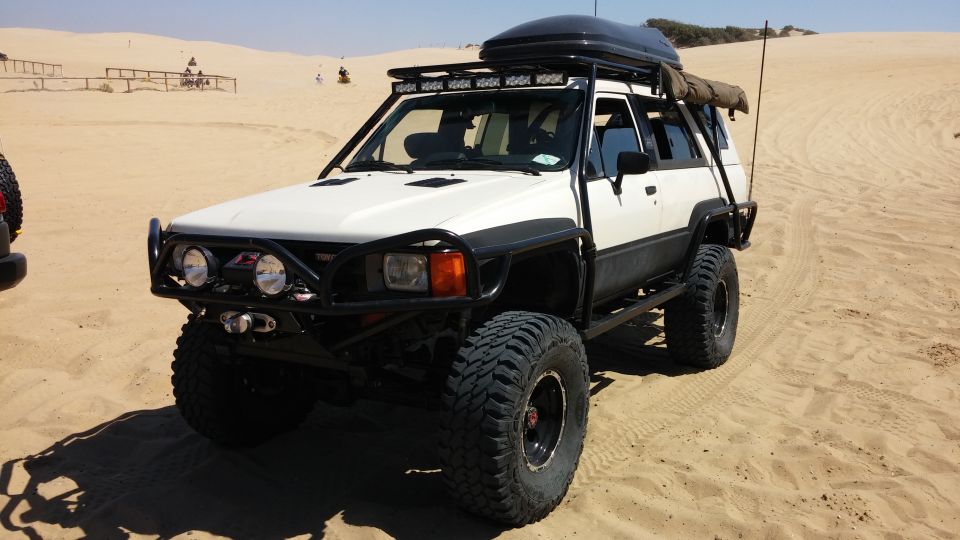 White Toyota 4Runner with a roof-top cargo box, light bar, and custom bumper, parked on sand dunes