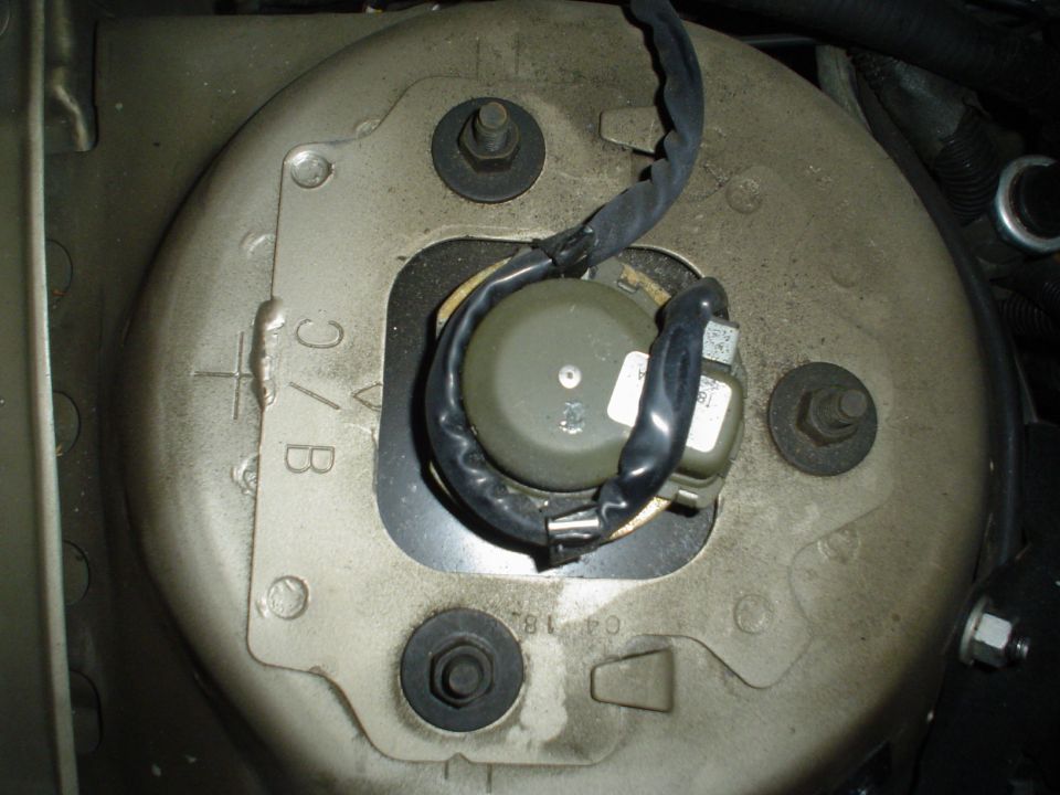 Top-down view of a suspension strut tower mount with three stud nuts and a wired sensor at center