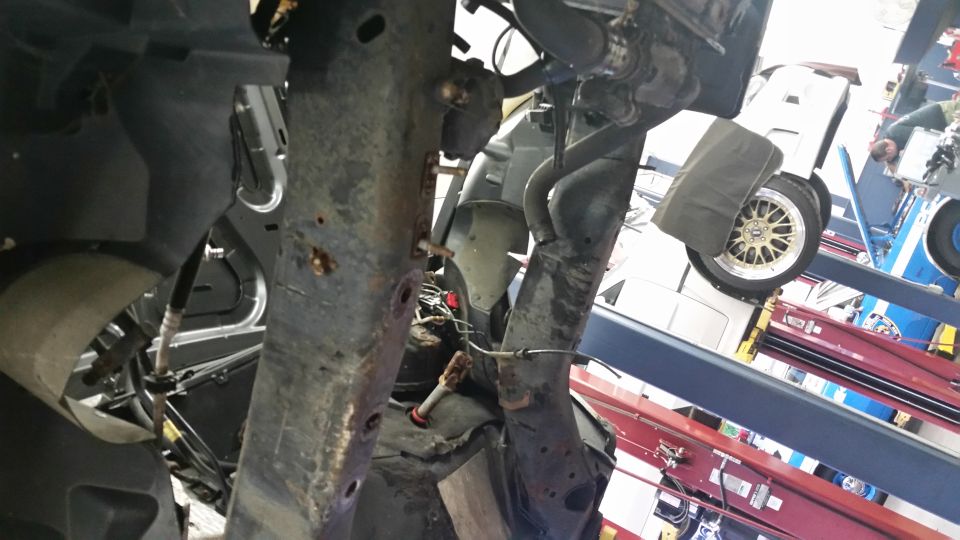 Rusted frame and suspension components of a vehicle, with a gold-spoked wheel visible in the background
