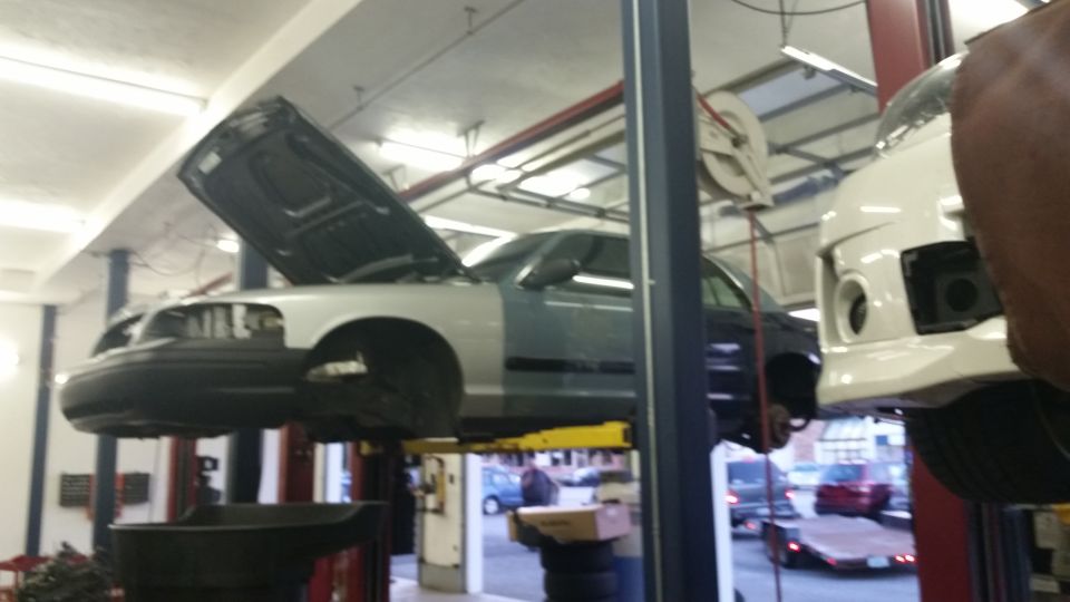 Silver and dark blue Ford Crown Victoria on a lift, hood open, front end partially disassembled