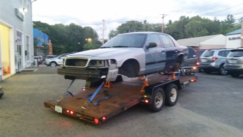 2001 Ford Crown Victoria - A 2001 Ford Crown Victoria, partially disassembled and loaded onto a trailer, is parked outside a building.