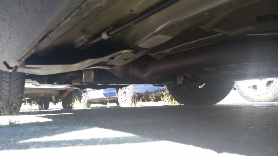 Underside of a car showing exhaust pipe and suspension components. Another vehicle is visible in the background
