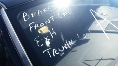 2001 Ford Crown Victoria - Chalk markings on a car's windshield list various parts and issues, including brakes, front end, exhaust, and trunk lock.