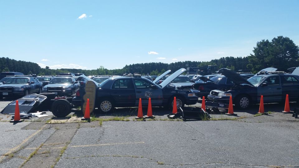 Several police cars parked in a lot, some with hoods open. Parts are scattered around
