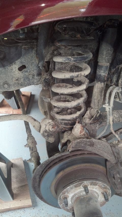 1996 Ford Bronco - This is a close-up, low-angle shot of the front suspension of a vehicle, likely a 1996 Ford Bronco, showing a heavily soiled coil spring, s…