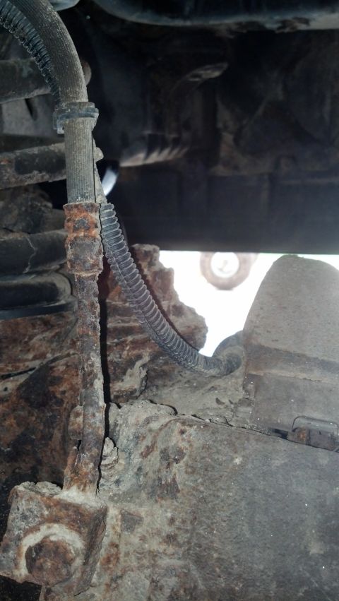 1996 Ford Bronco - This image shows a heavily rusted brake line and suspension components on the underside of a vehicle. A black protective sleeve covers part…