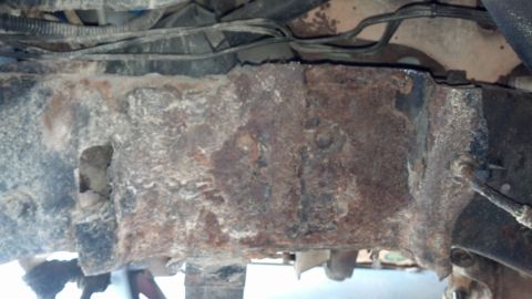 1996 Ford Bronco - The underside of a vehicle shows significant rust and corrosion on the frame and associated components. Wires and cables are visible above…