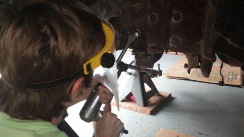 1996 Ford Bronco - A person wearing a face shield and light green shirt uses a tool to work on the rusty undercarriage of a vehicle, which is supported by jac…