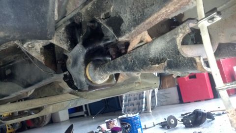 1996 Ford Bronco - The underside of a vehicle shows a heavily rusted and possibly damaged suspension component, with a yellow bushing visible. Tools and stora…