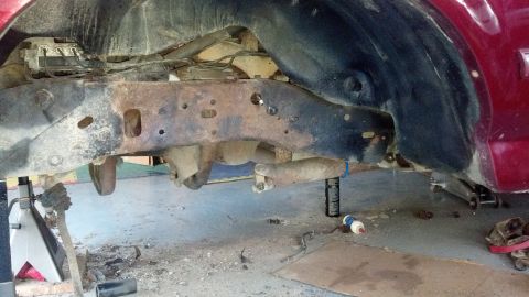 1996 Ford Bronco - A close-up, low-angle view of the heavily rusted frame and undercarriage of a red SUV, possibly a 1996 Ford Bronco, with various suspension…