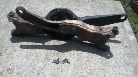 1996 Ford Bronco - A rusty differential housing and control arm assembly are laid out on a concrete surface, with two small metal brackets in the foreground.
