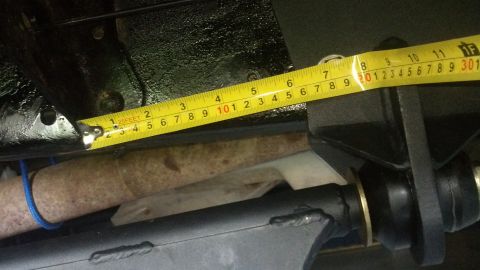 1996 Ford Bronco - A yellow measuring tape is shown extended across a rusty exhaust pipe and black suspension components. The tape measures approximately 8 in…