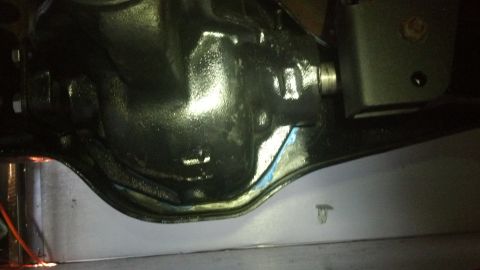 1996 Ford Bronco - A close-up view of a black differential housing with a light blue sealant applied around the gasket area. A silver metal shield is visible…