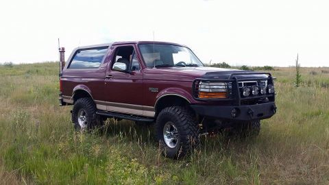 1996 Ford Bronco - A maroon 1996 Ford Bronco with a tan stripe is parked in a grassy field under a cloudy sky. The SUV features a black aftermarket front bump…