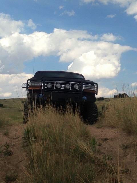 1996 Ford Bronco - A black Ford Bronco is parked on a dirt path in a grassy, hilly area under a cloudy blue sky. The vehicle has a prominent grille guard with…