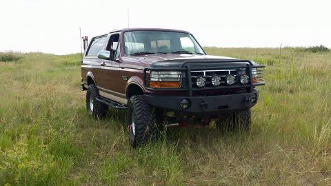 1996 Ford Bronco - A maroon 1996 Ford Bronco is parked in a grassy field. The SUV features a black brush guard with four KC lights and a heavy-duty aftermarke…