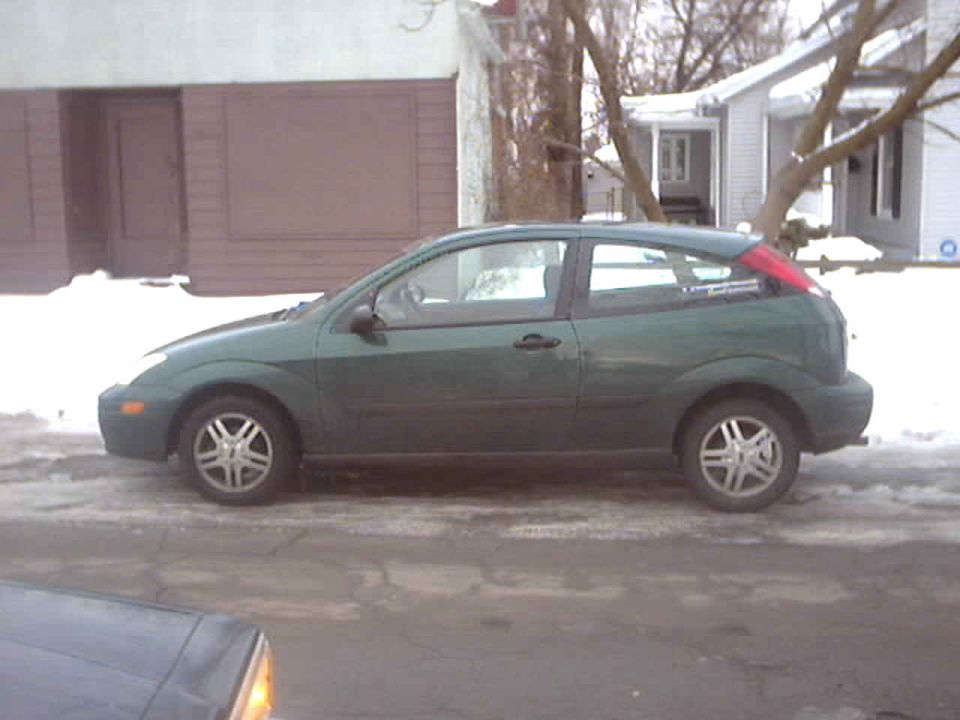 Dark green Ford Focus 3-door hatchback, side view, parked on a snowy street in front of a garage
