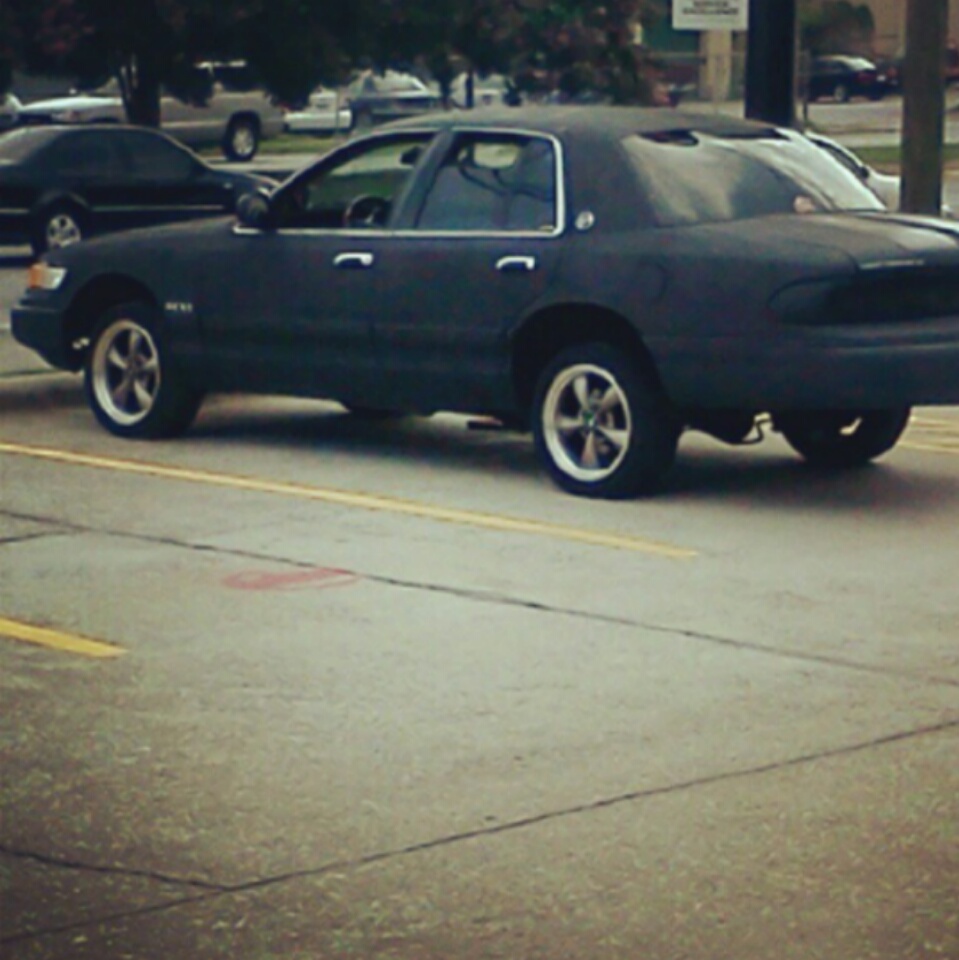 Dark matte full-size sedan with vinyl roof and chrome wheels parked street-side, side rear view.