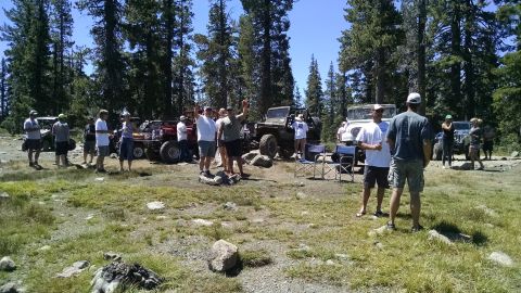 1985 Jeep CJ7 - A group of people stand around several off-road vehicles, including a red Jeep, in a wooded, grassy area on a sunny day. One person holds u…