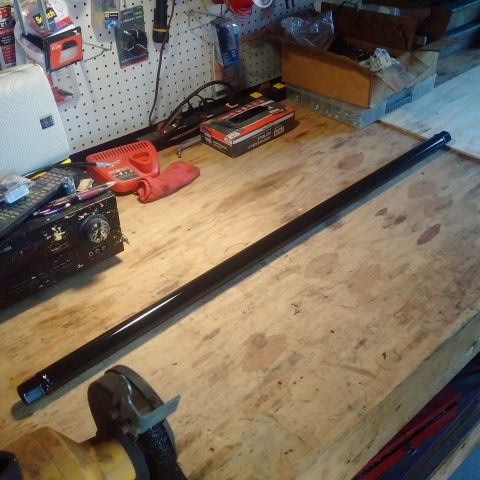 1978 Ford Bronco - A long, black, cylindrical part, possibly a drive shaft or exhaust pipe, lies on a wooden workbench. Various tools and supplies are visible…