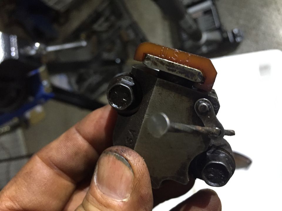 Hand holding a timing chain tensioner with orange U.S.A.-marked plastic guide shoe and two bolts
