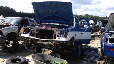 1994 Ford Bronco - A blue and white GMC truck sits in a junkyard with its hood open, showing missing parts.
